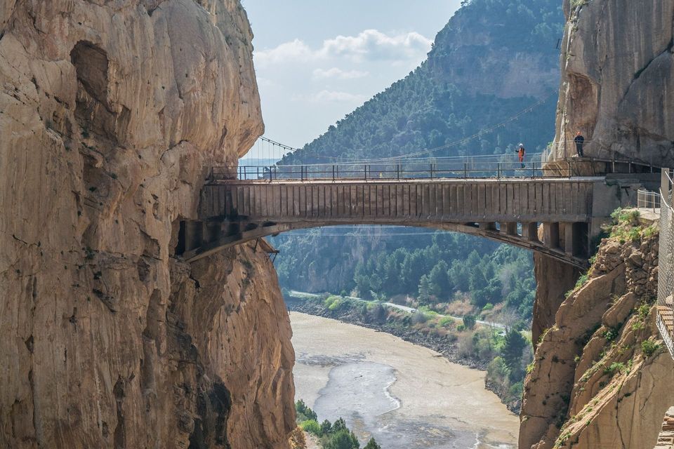 Un ponte alto collega due ripide pareti rocciose su un ampio fiume in un canyon profondo, con due persone in piedi.