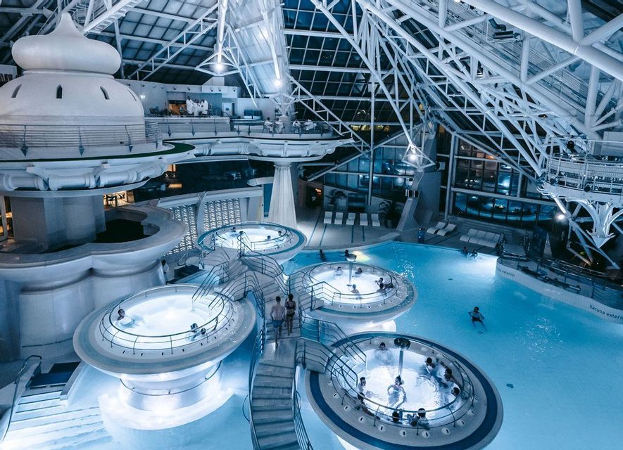 Ci si rilassa in diverse piscine circolari illuminate all'interno di una spa termale multilivello con un ampio soffitto geometrico bianco.