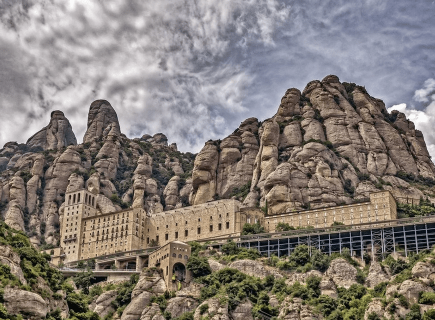 Un grande monastero di pietra incastonato nel fianco di una ripida montagna rocciosa sotto un cielo drammatico e nuvoloso.