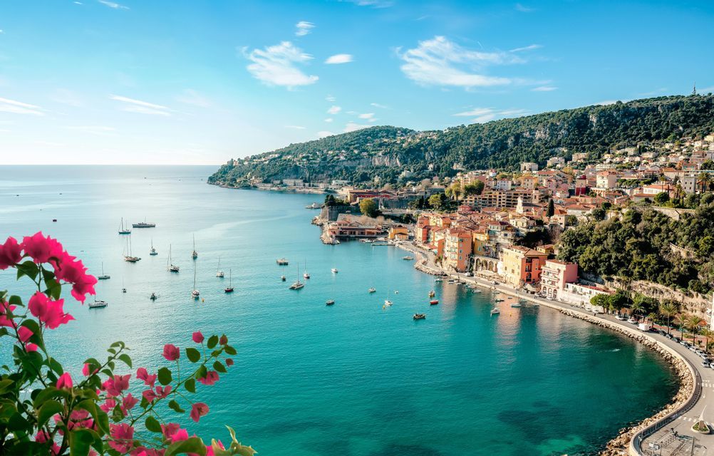 Une ville côtière colorée sur une colline verdoyante surplombant une baie turquoise avec des voiliers, encadrée par des fleurs roses au premier plan.