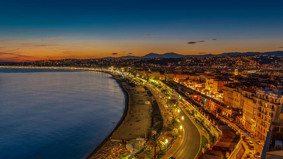 Une ville côtière illuminée au crépuscule, avec une promenade incurvée longeant la plage et la mer, et des montagnes visibles au loin.