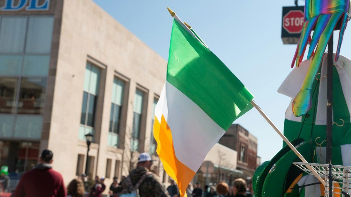Gros plan sur un drapeau irlandais flottant au soleil lors d'une parade de rue, avec des bâtiments de la ville en arrière-plan.