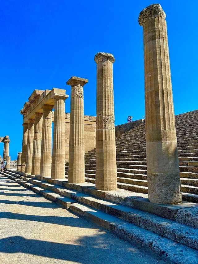 Alte colonne in pietra di un'antica rovina proiettano lunghe ombre accanto a un'ampia scalinata sotto un cielo azzurro vibrante.