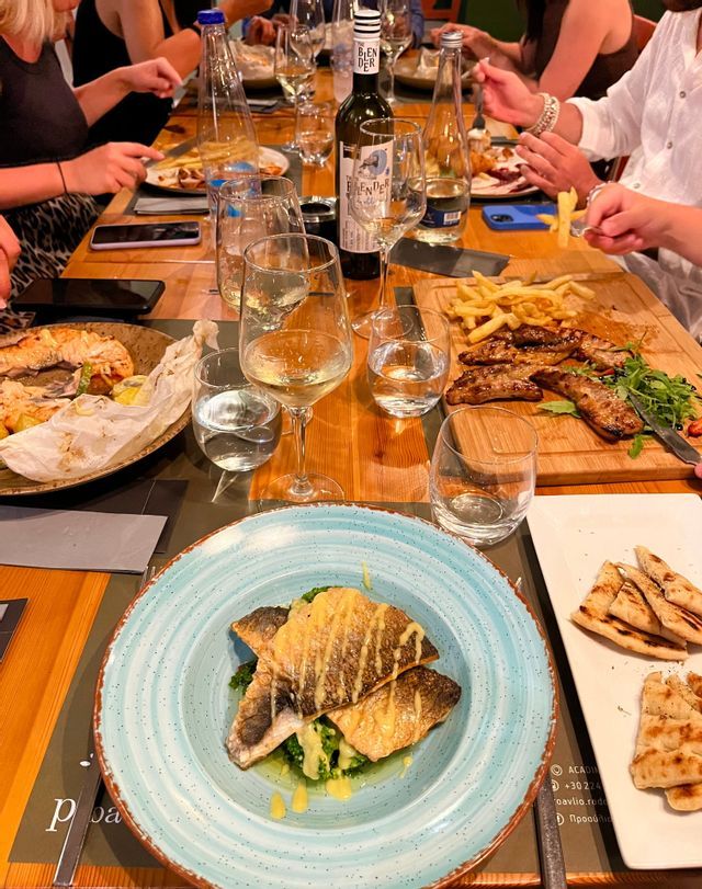 Un viaggio di gruppo WeRoad a cena a un lungo tavolo di legno, imbandito con piatti di pesce alla griglia, carne, patatine fritte e bicchieri di vino bianco.