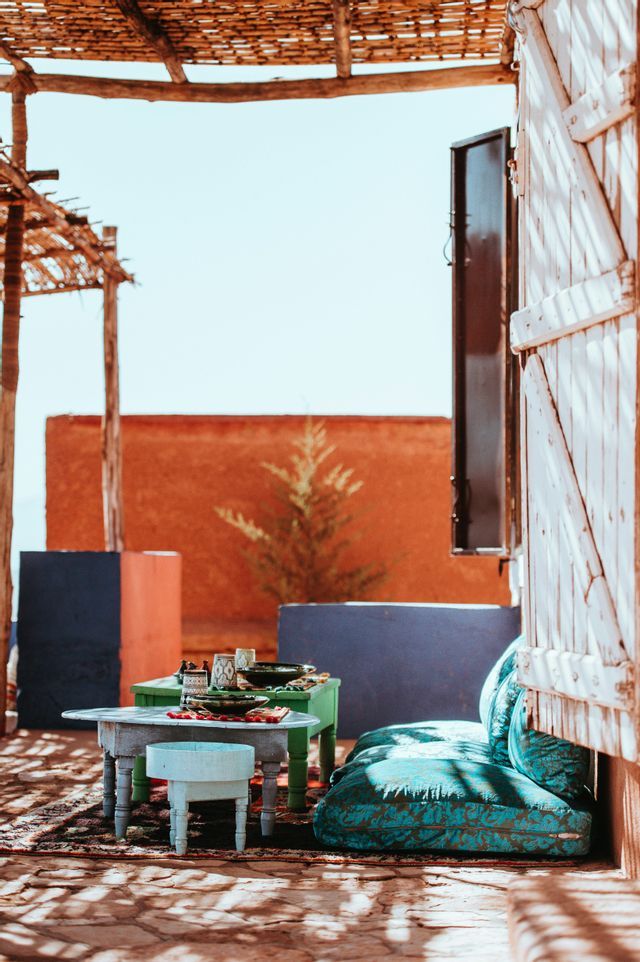 Un salon de patio extérieur rustique avec des tables basses, des coussins de sol turquoises et un toit de roseaux projetant des ombres par une journée ensoleillée.