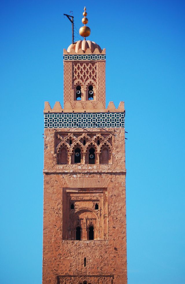 Un minaret en pierre orné avec des sculptures complexes, des carreaux décoratifs et un fleuron doré, se détachant sur un ciel bleu clair.