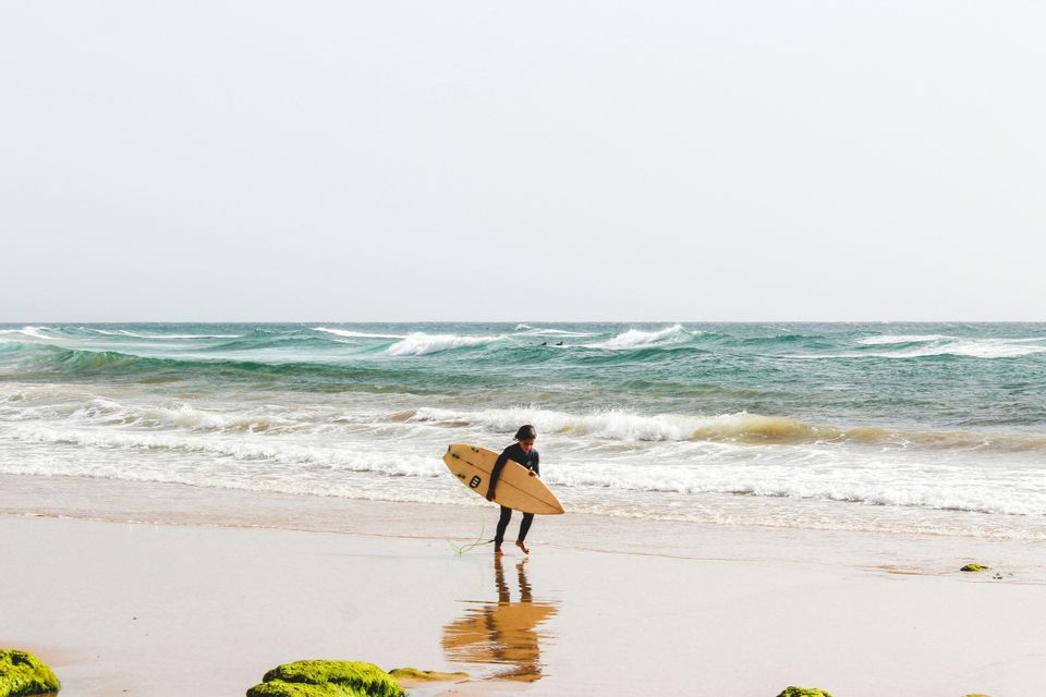 Un surfeur en combinaison noire porte une planche de surf en marchant sur le sable mouillé d'une plage avec des vagues en arrière-plan.