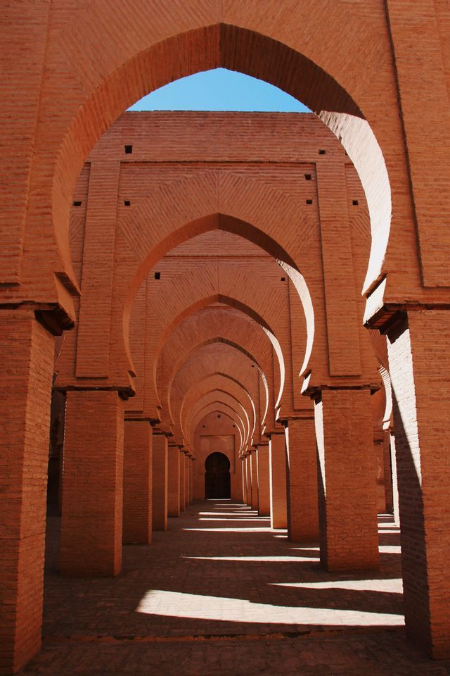 Un long couloir en briques rouges avec des arches et des colonnes répétées, où la lumière du soleil projette de fortes ombres sur le sol.