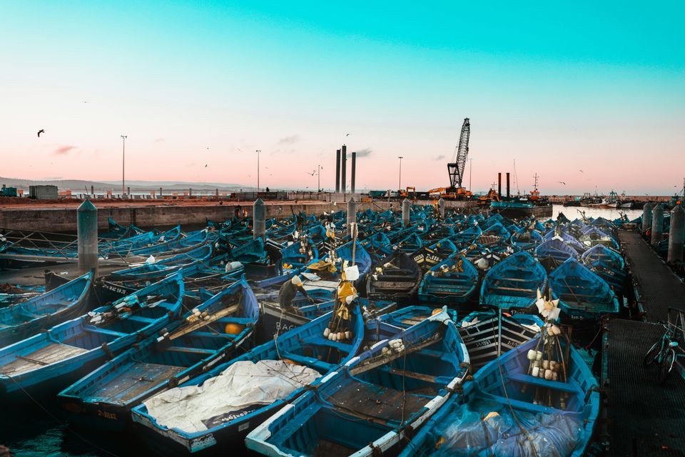 Une flotte de petits bateaux de pêche bleus est amarrée dans un port animé sous un ciel rose et turquoise.