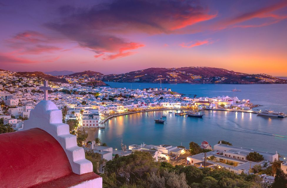 Una chiesa dalla cupola rossa domina una cittadina costiera illuminata, con edifici imbiancati attorno a un porto al tramonto.