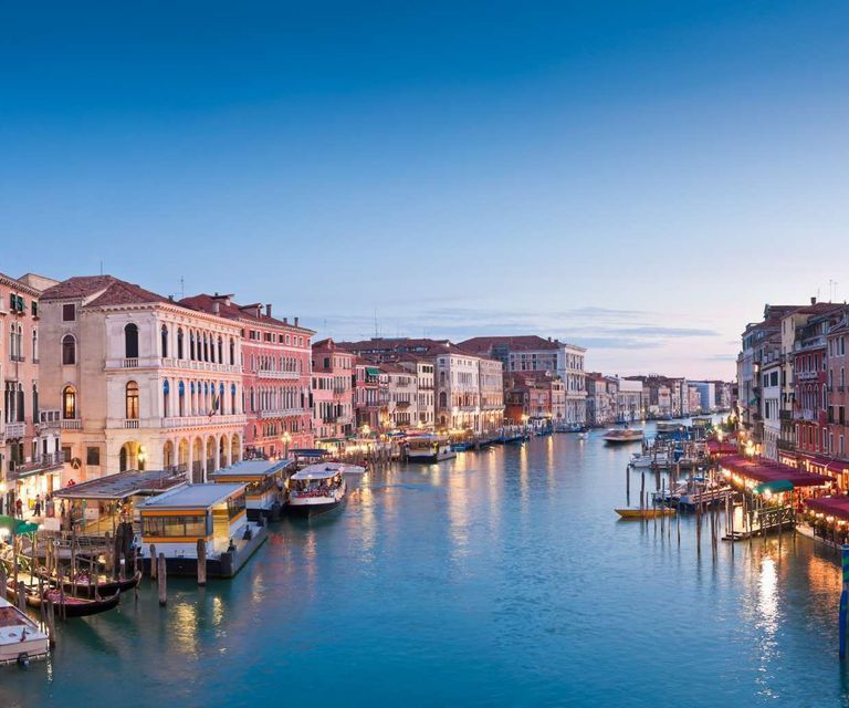 Edifici storici illuminati fiancheggiano un ampio canale pieno di barche e vaporetti sotto un cielo azzurro e limpido al tramonto.