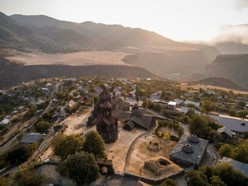 Un antico monastero in pietra e un villaggio circostante su una collina soleggiata con montagne sfocate sullo sfondo.