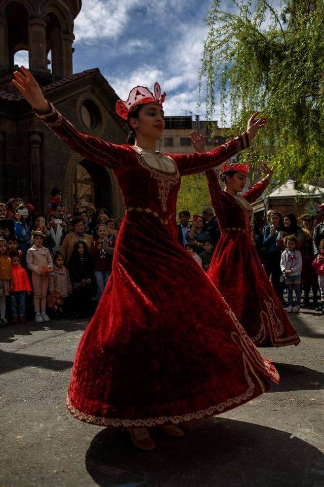 Due donne, in abiti tradizionali di velluto rosso e copricapi, si esibiscono in una danza per il pubblico all'aperto, di fronte a un edificio in pietra.