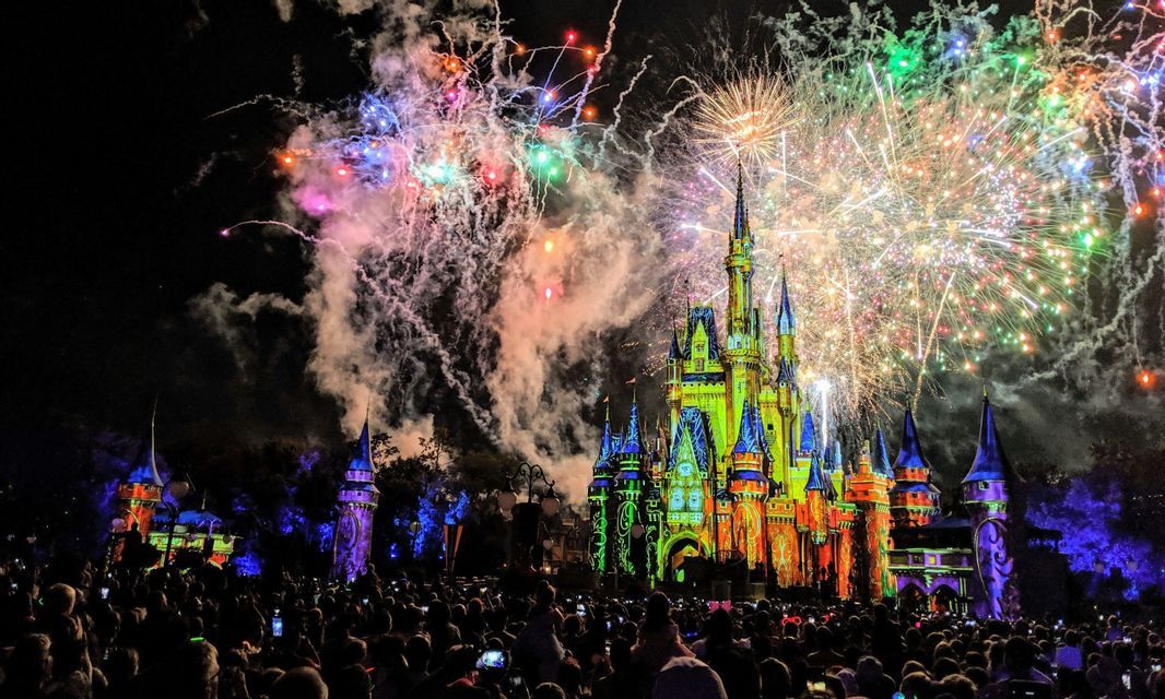 Colorful fireworks explode over a brightly illuminated fairytale castle as a large crowd watches the show at night.