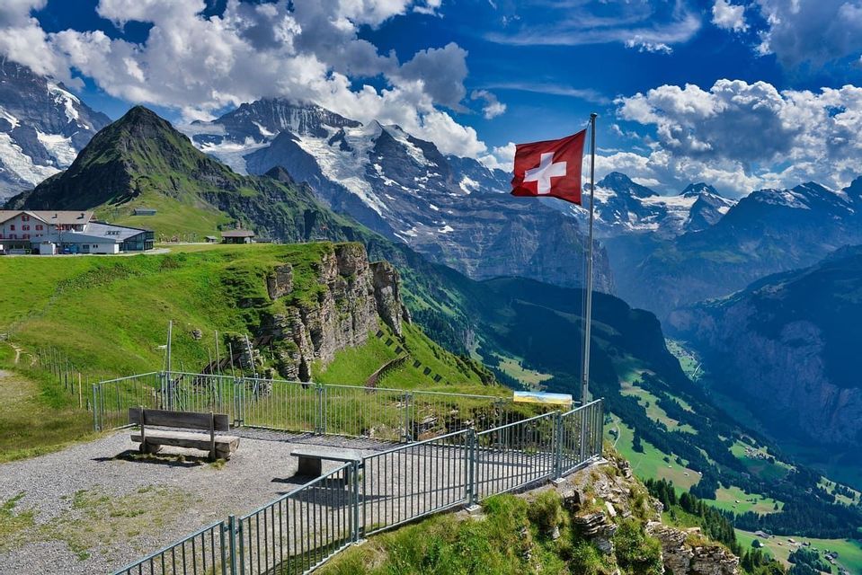 Un drapeau suisse sur un mât se dresse à un point de vue dominant une vallée verdoyante et une chaîne de montagnes enneigées.