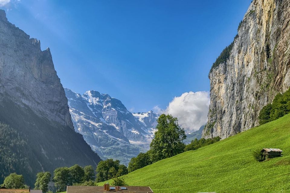 Une vue sur une vallée verdoyante encadrée par des falaises abruptes, avec des montagnes enneigées en arrière-plan sous un ciel bleu.