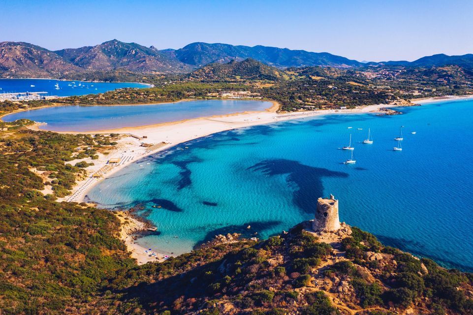 An aerial view of a stone tower on a green hill overlooking a white sand beach, a lagoon, and turquoise water with sailboats.