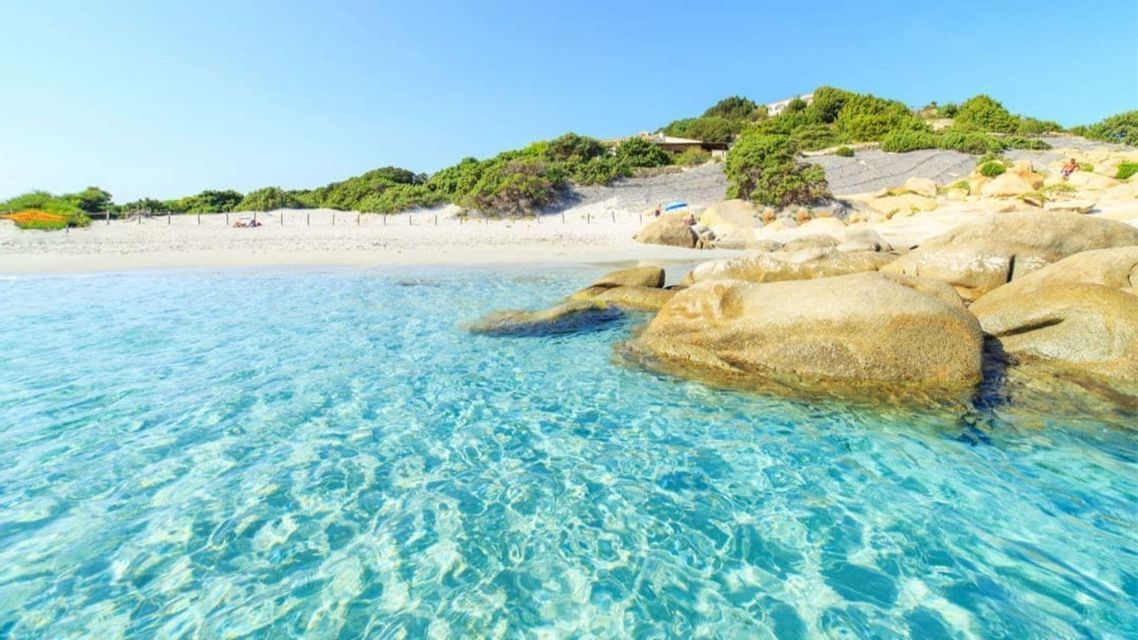 Clear turquoise water flows around large rocks towards a white sand beach with a green hill in the background.