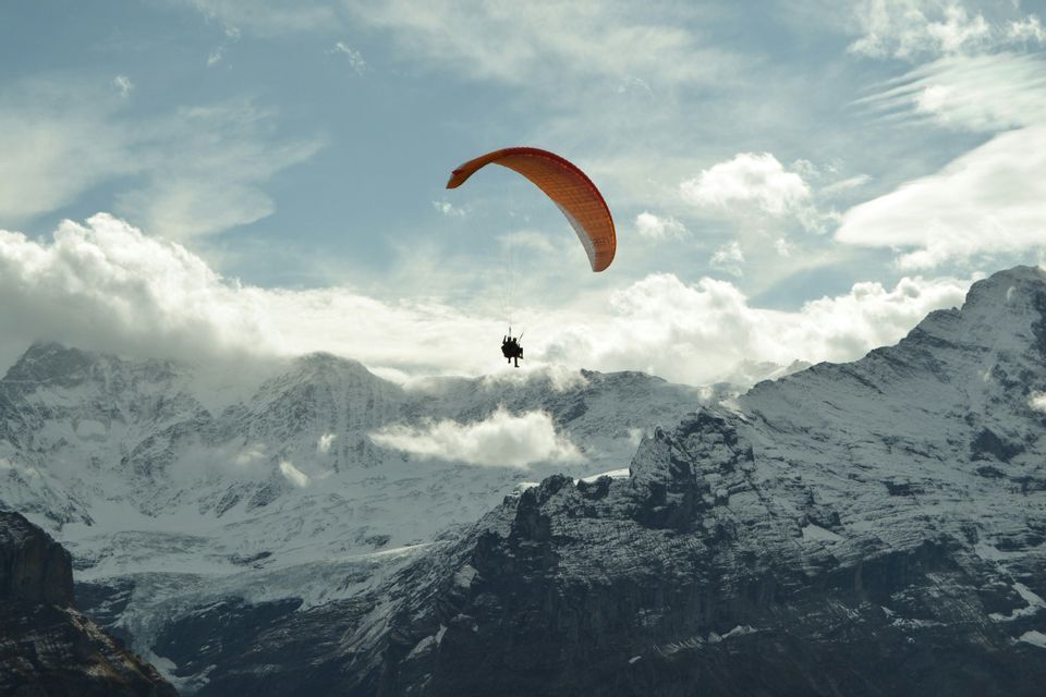 Deux personnes en parapente biplace avec une aile orange s'envolent au-dessus d'une vaste chaîne de montagnes enneigée sous un ciel nuageux.
