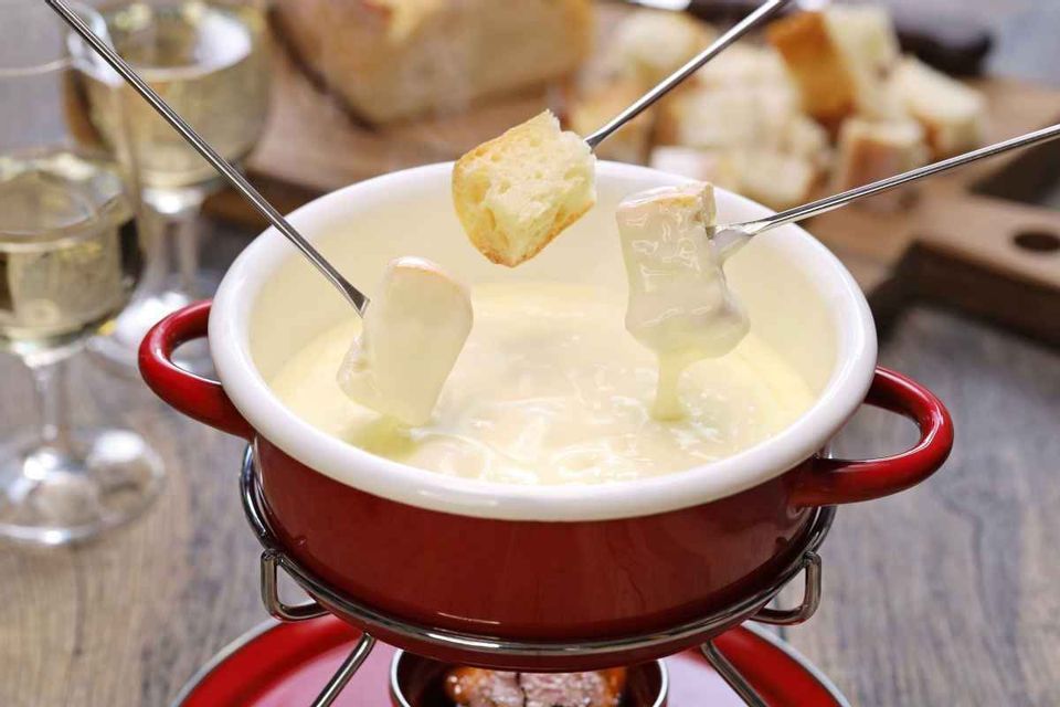 Cubes of bread on three fondue forks are dipped into a red pot of melted cheese fondue on a wooden table.