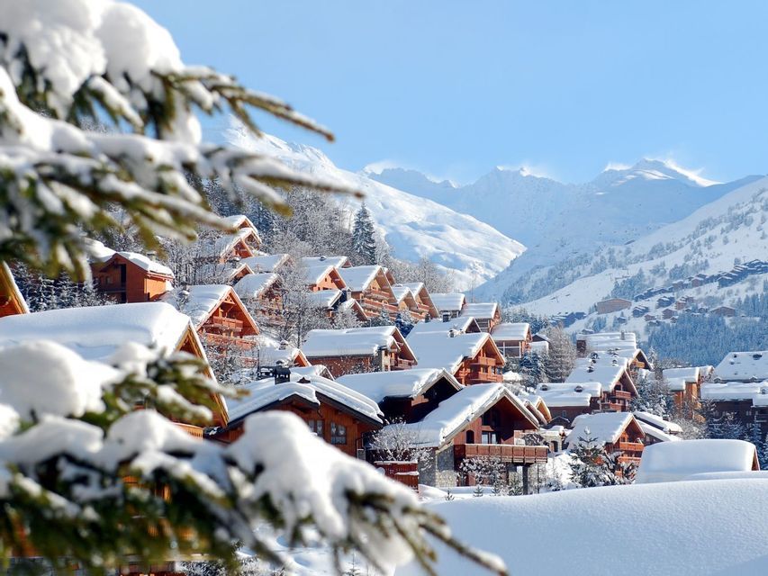 Un gruppo di chalet in legno coperti da neve alta su un fianco di montagna, con cime innevate sullo sfondo.