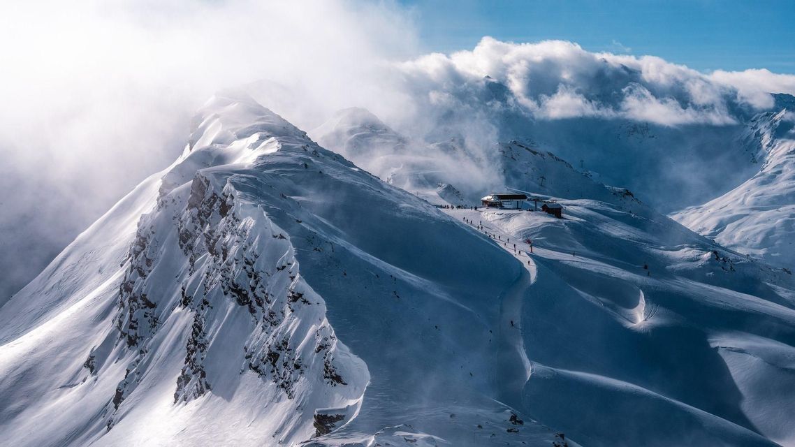 A vast, snow-covered mountain range on a windy day, with skiers descending a slope past a mountain lodge.