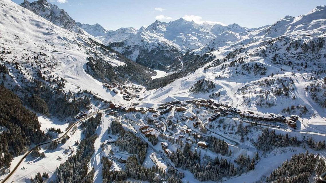 An aerial view of a ski resort village nestled in a snowy valley surrounded by mountain peaks and pine forests.
