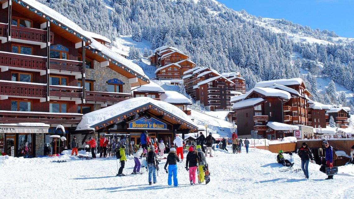 Una vivace località sciistica in una giornata di sole, con persone in attrezzatura invernale che sciano e fanno snowboard vicino a grandi chalet di legno coperti di neve.