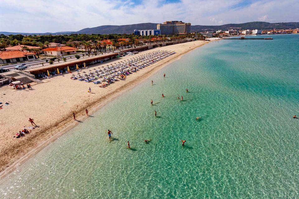 Vista aerea di una spiaggia soleggiata con persone che nuotano in acque limpide e turchesi e si rilassano sotto file di ombrelloni.