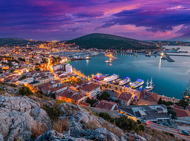 Una città costiera e il suo porto illuminati dalle luci, visti da un punto panoramico roccioso al crepuscolo sotto un cielo viola.