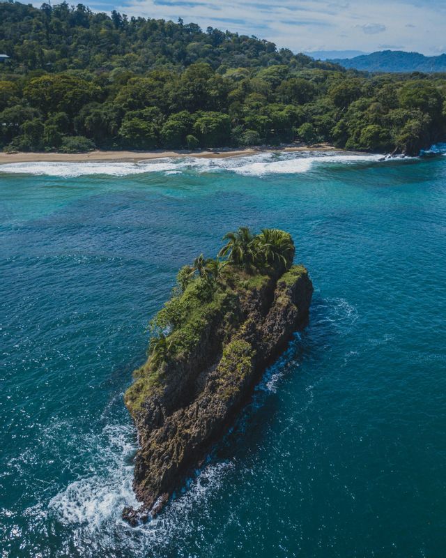 Una vista aerea di un isolotto roccioso con palme nell'oceano turchese, con una costa boscosa sullo sfondo.