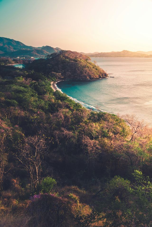 Veduta dall'alto di una costa tropicale con baia isolata, acqua turchese e lussureggianti colline verdi sotto un cielo velato e illuminato dal sole.
