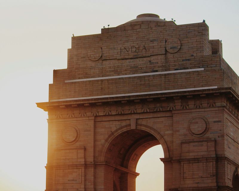 Un primer plano de un gran arco de piedra con "INDIA" inscrito en su fachada, iluminado por el sol dorado contra un cielo brillante.
