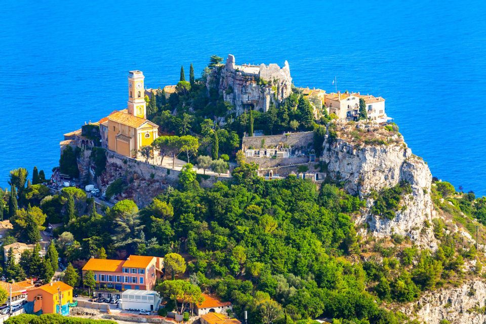 Un village dominé par son clocher est perché sur une falaise rocheuse luxuriante et arborée, surplombant une vaste mer bleue.