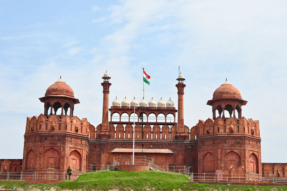 La bandera india ondea sobre un gran fuerte de arenisca roja con intrincadas torres y cúpulas bajo un cielo azul claro.