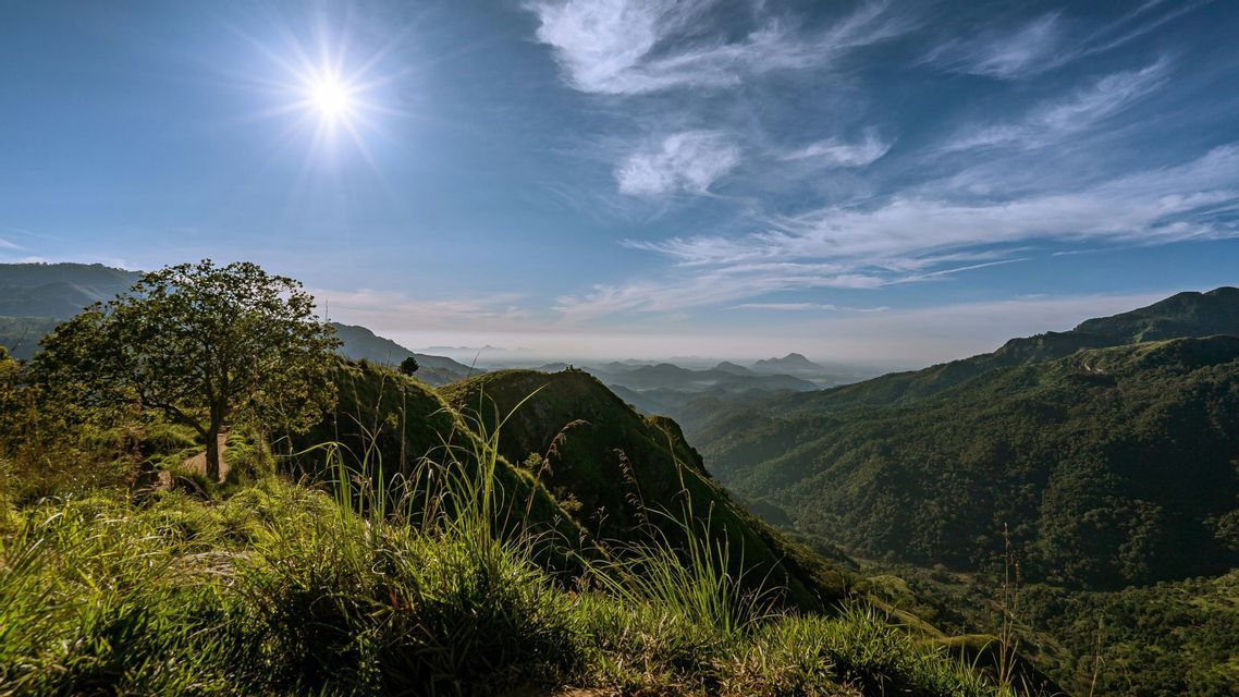 Un sol radiante ilumina un vasto paisaje de colinas y valles verdes ondulantes, visto desde una ladera cubierta de hierba bajo un cielo azul.