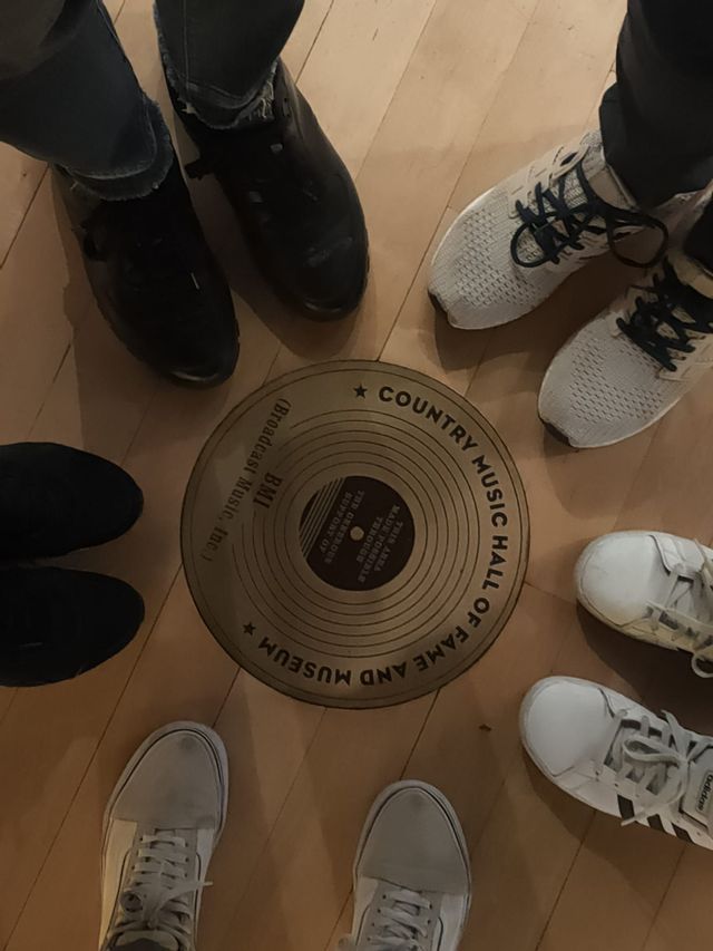 Una veduta dall'alto dei piedi di un gruppo WeRoad, in piedi attorno a una targa a pavimento della Country Music Hall of Fame e del Museo.
