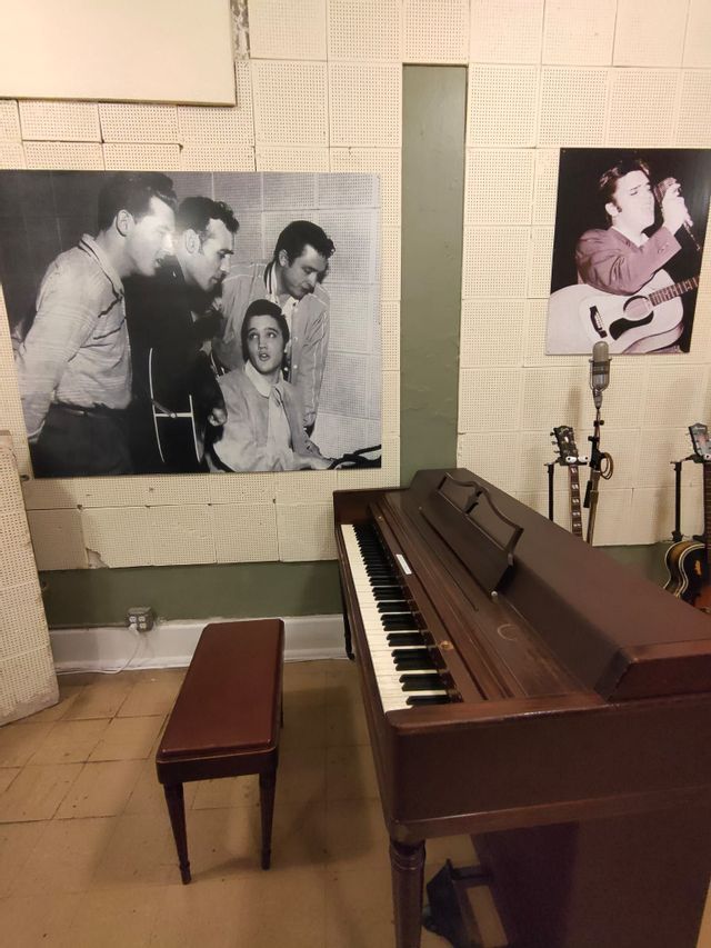 Uno studio musicale con un pianoforte verticale, una panca, microfoni vintage e grandi fotografie di Elvis Presley alla parete.
