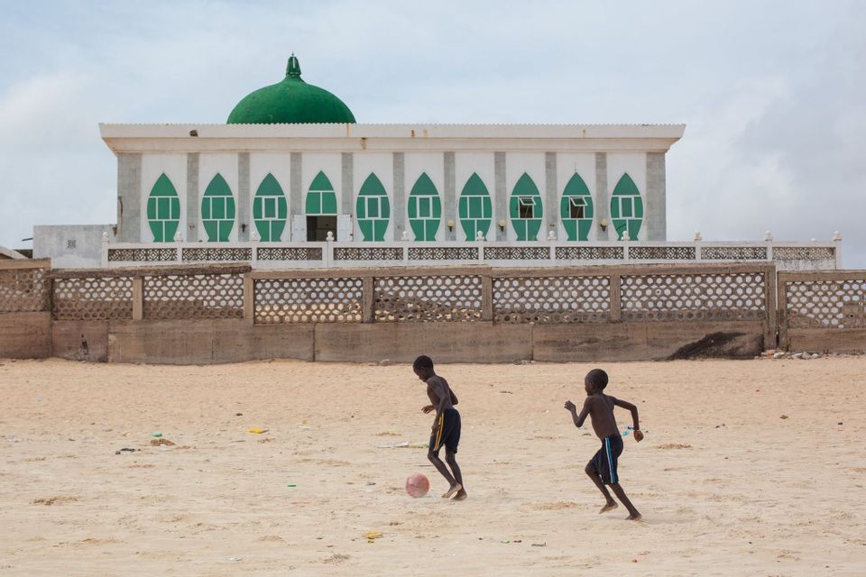 Due giovani ragazzi giocano a calcio su un terreno sabbioso davanti a una grande moschea bianca e verde con una cupola.