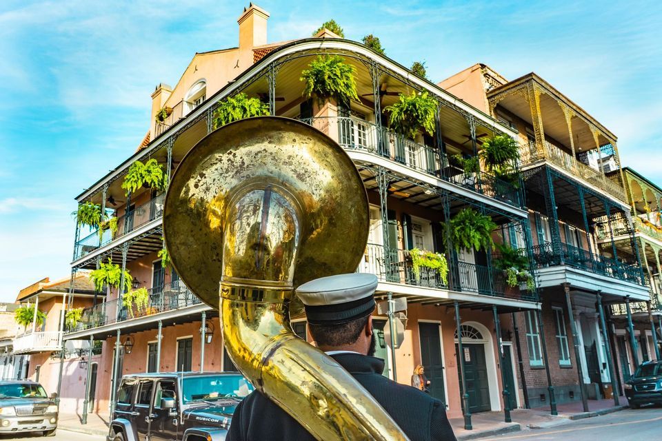 Un musicista visto di spalle porta una tuba lungo una strada fiancheggiata da edifici con balconi in ferro battuto ornati.