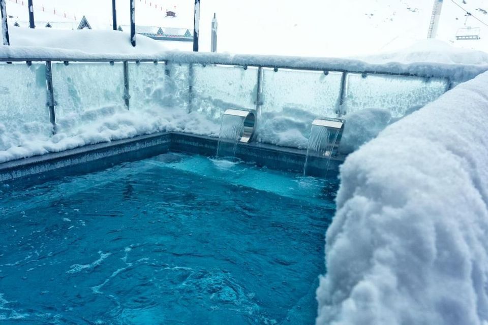 Una vasca idromassaggio all'aperto con due getti a cascata che si riversano nell'acqua azzurra, circondata da neve alta in un resort di montagna.