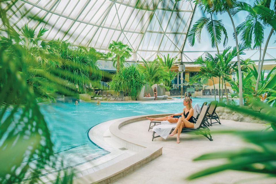 Una donna in costume si rilassa su una sdraio a bordo di una piscina coperta circondata da piante tropicali sotto una cupola di vetro.