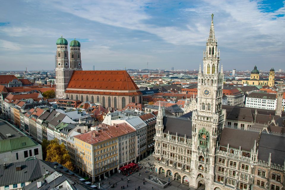 Veduta aerea di una storica città europea con un grande municipio gotico e una cattedrale con due torri a cupola verde sotto un cielo azzurro.