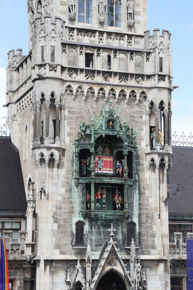 Un carillon verde riccamente decorato con figure colorate, incastonato nella facciata in pietra di una torre dell'orologio gotica.
