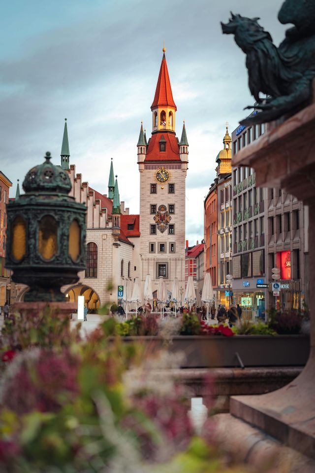 Una storica torre dell'orologio con un tetto rosso appuntito si erge su una piazza europea, vista oltre fiori sfocati al tramonto.
