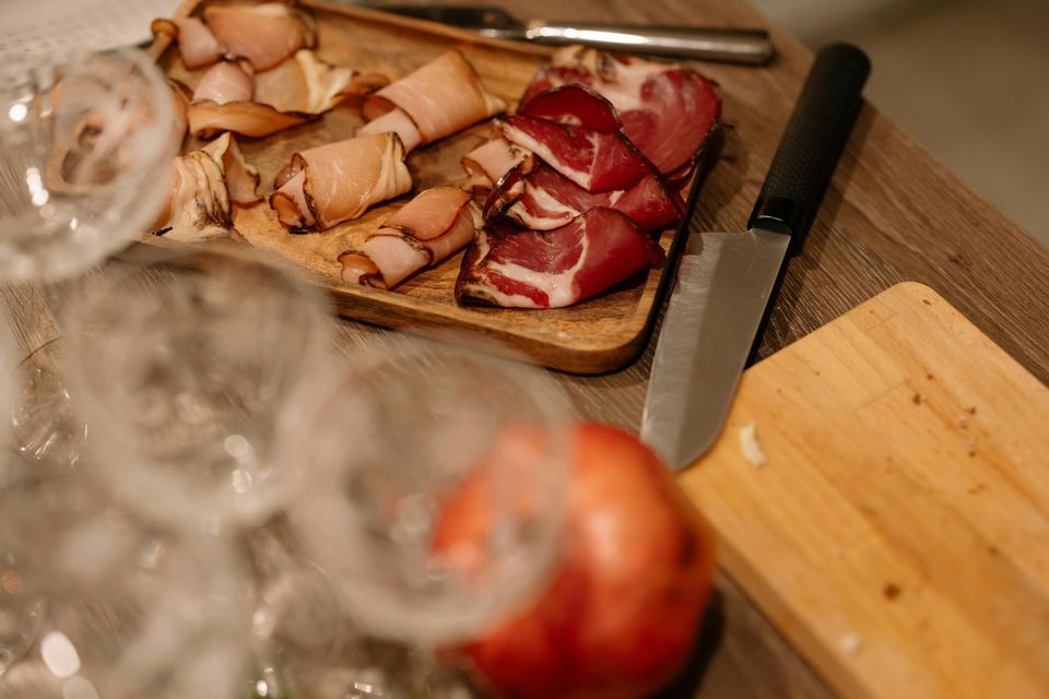 Un tagliere di legno con vari salumi affettati e arrotolati si trova accanto a un tagliere e un coltello, con bicchieri sfocati in primo piano.