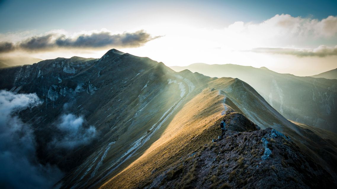 Un escursionista con uno zaino cammina lungo una stretta cresta di montagna mentre i raggi del sole illuminano il paesaggio all'alba.