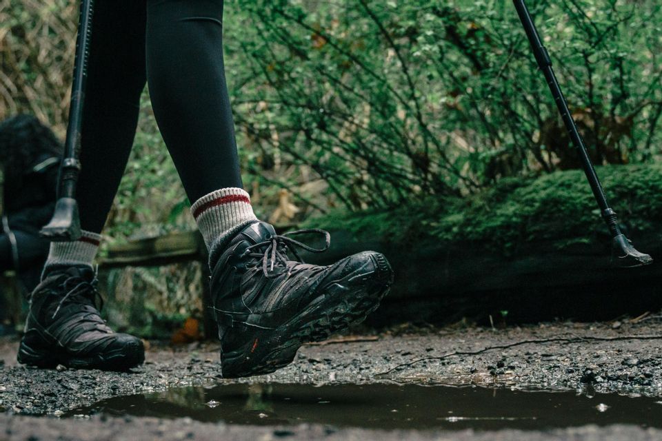 Primo piano di scarponi e calzini da trekking mentre una persona scavalca una pozzanghera su un sentiero sterrato in un bosco.