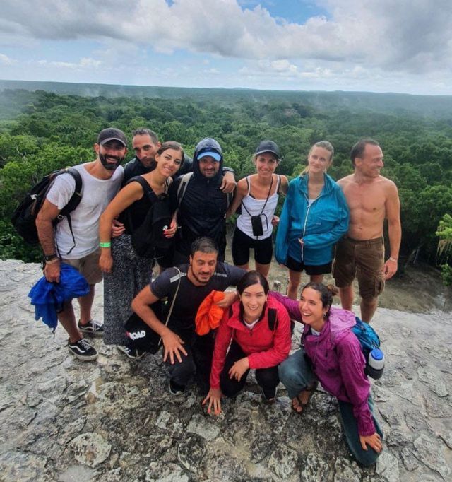 Un gruppo WeRoad in viaggio posa per una foto su un punto panoramico roccioso, con una vasta foresta verde che si estende sotto un cielo nuvoloso.