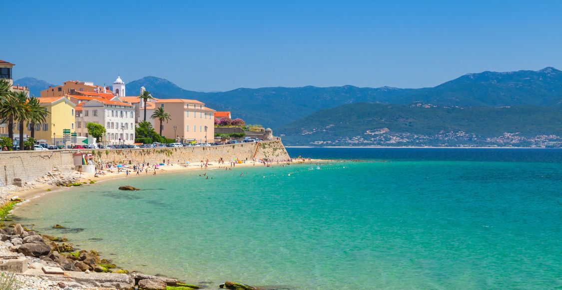 Une ville côtière avec une digue en pierre à côté d'une plage de sable où les gens nagent dans l'eau turquoise sous un ciel bleu clair.
